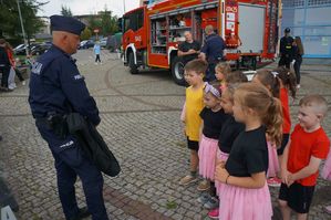 na zdjęciu policjant stoi przed dziećmi, w tle wóz strażacki